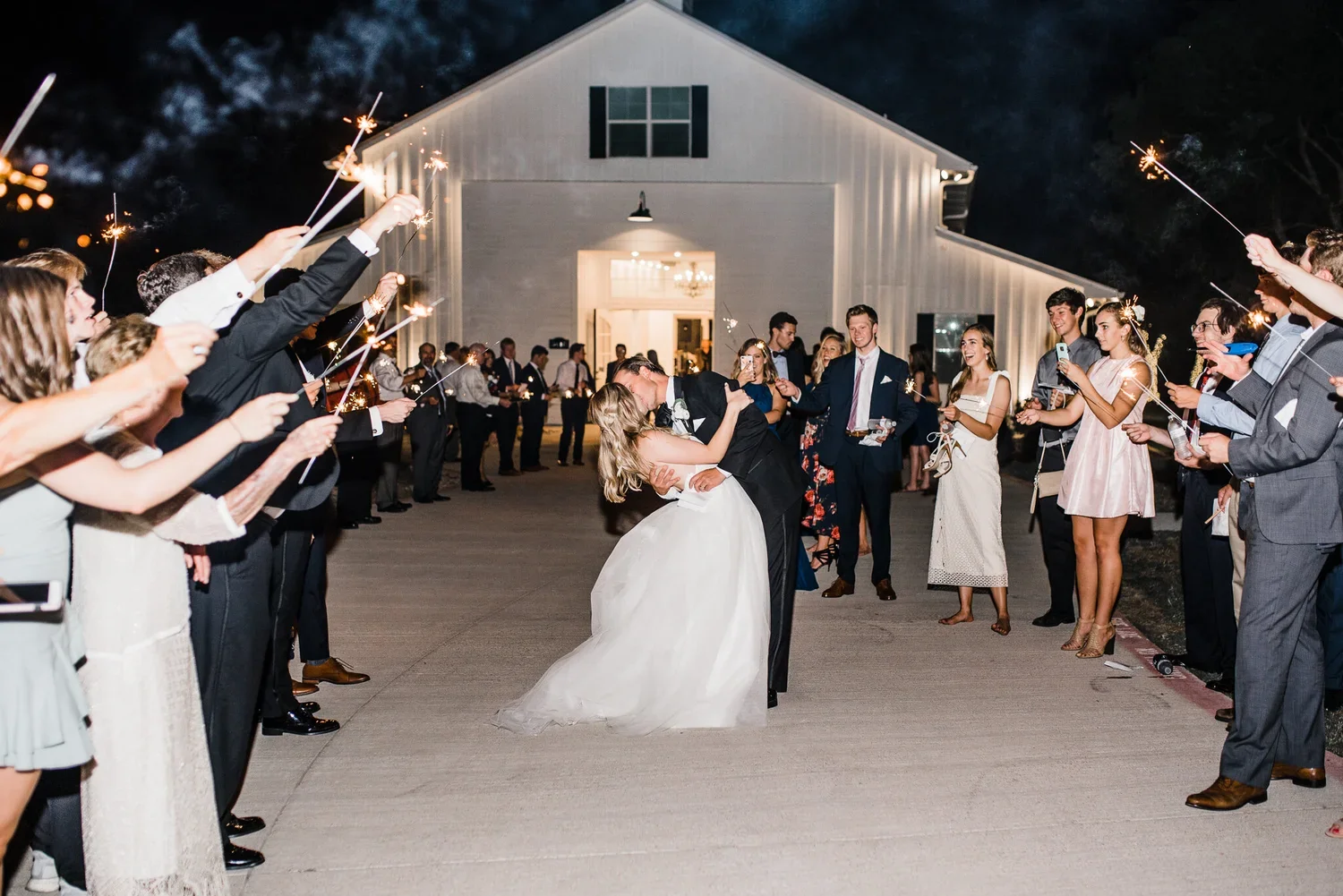 A bride and groom kiss in the center of a large outdoor gathering holding sparklers at night, with a barn in the background and guests on both sides.