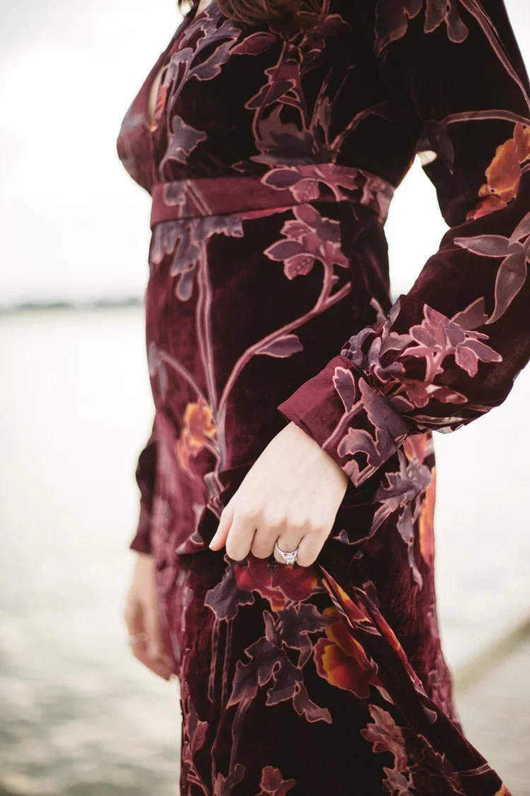 Close-up of a woman in a floral velvet dress with a ring on her finger, standing outdoors near a body of water.