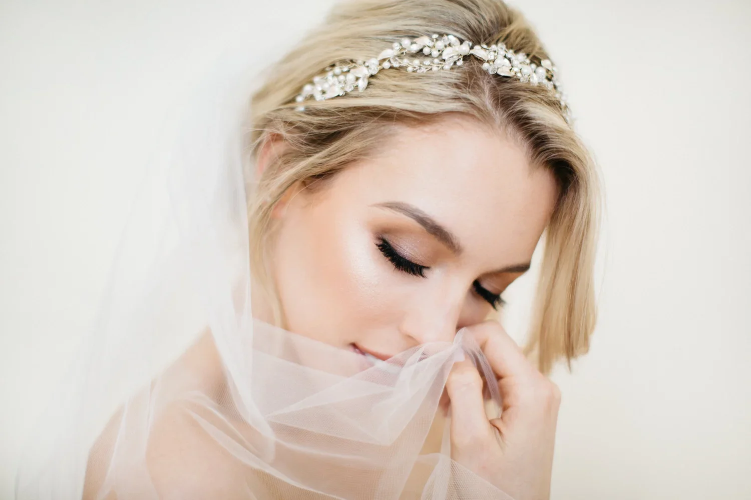 A bride with blonde hair styled with a bejeweled headband, holding a sheer wedding veil near her face with eyes closed, makeup, and a serene expression.