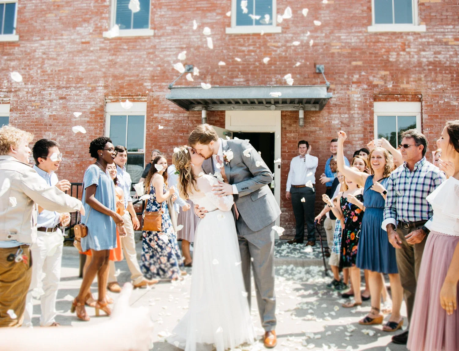 Bride and groom kissing while guests celebrate with flower petals outside a brick building at a wedding celebration.
