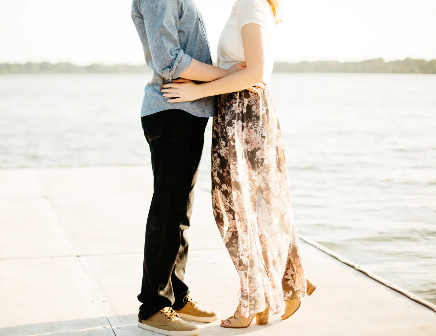A couple standing close together by the water, holding each other with their hands on each other's waists.
