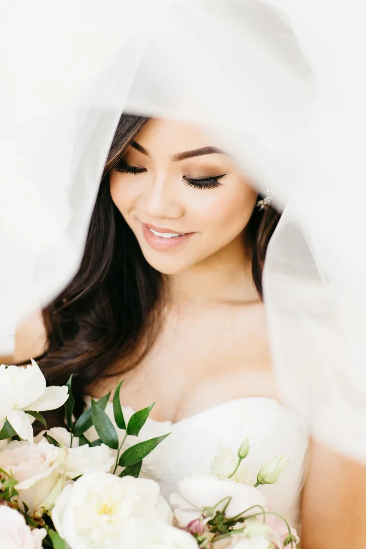 A woman in a wedding dress, smiling and looking down at a bouquet of white flowers, partly covered by a white veil.