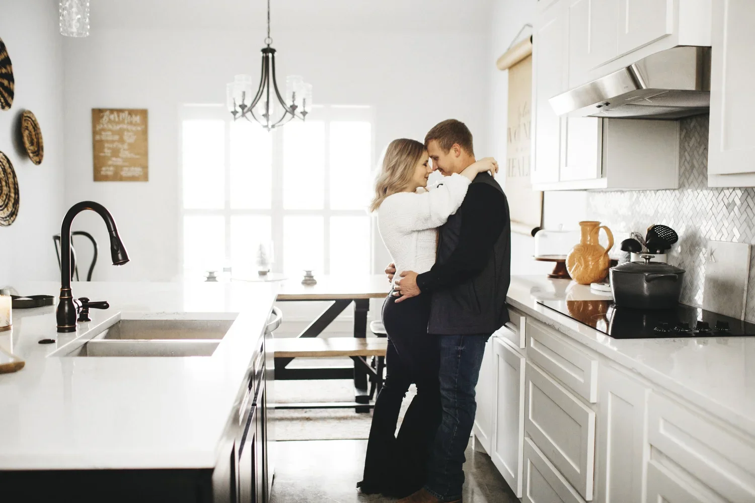 A couple embracing in a bright, modern kitchen with white cabinets, a kitchen island, and large windows.