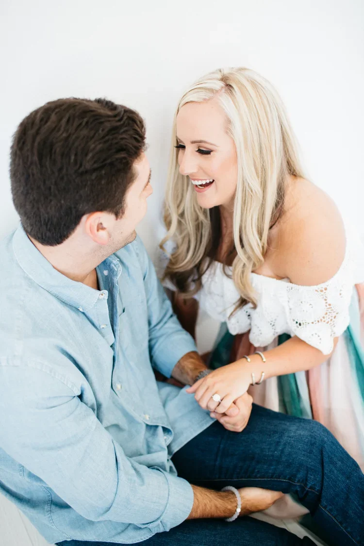 A happy woman with blonde hair, wearing a white off-the-shoulder top, holding hands with a man in a light blue shirt, both smiling and looking at each other.