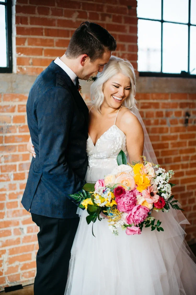 A bride and groom sharing a joyful moment indoors with a brick wall and large windows in the background. The bride is holding a colorful bouquet and is smiling, while the groom leans in toward her.