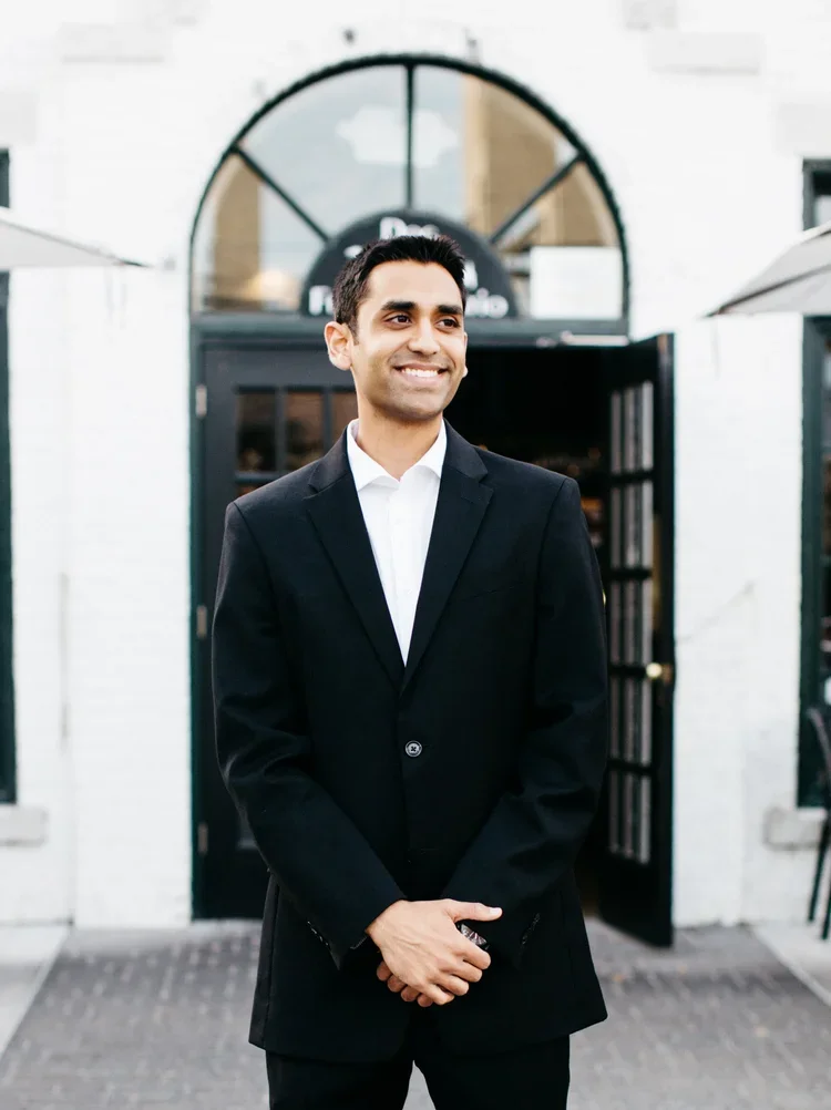 A man in a black suit and white shirt smiling outdoors in front of a building with a large arched window and open door.