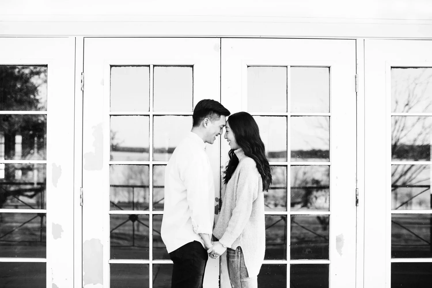 A black and white photo of a couple standing close together, holding hands, with their foreheads touching and smiling, in front of glass double doors with grid windowpanes.