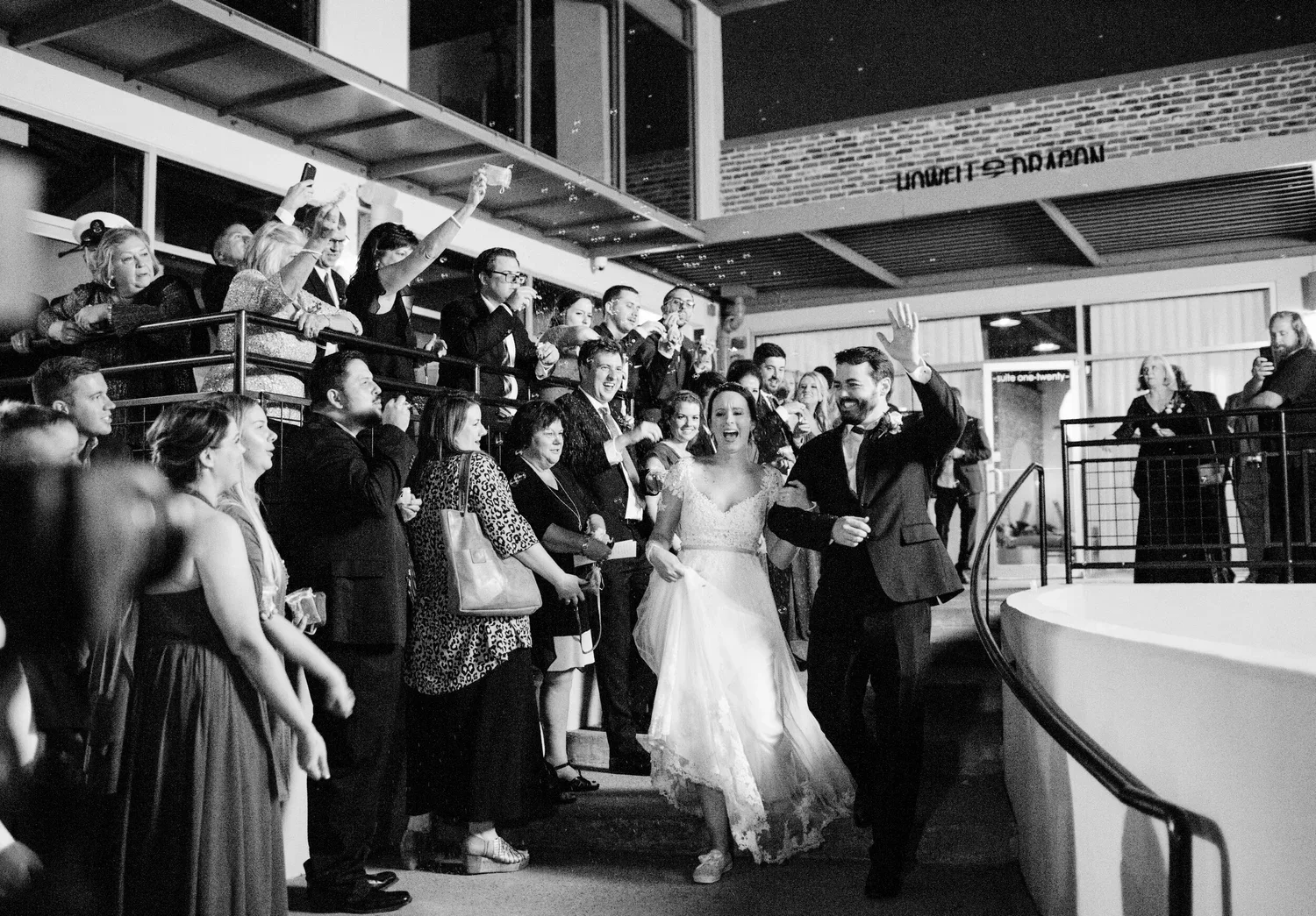 Black-and-white photo of a wedding celebration with a bride and groom walking down steps, surrounded by guests cheering and taking photos indoors.