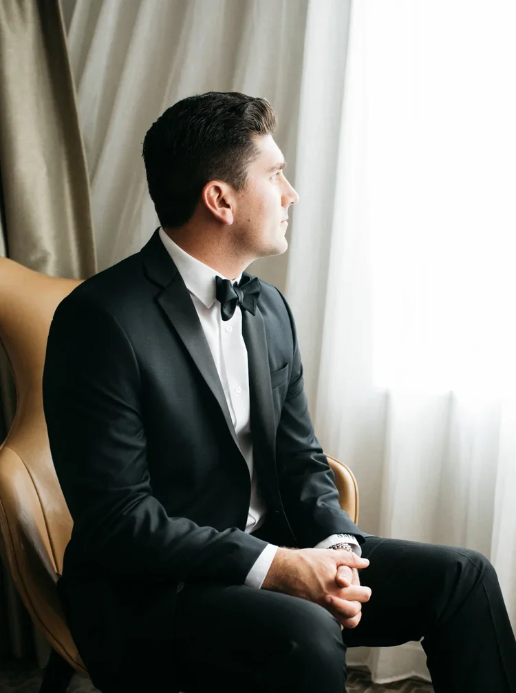 A man in a tuxedo sitting on a chair, looking out the window.
