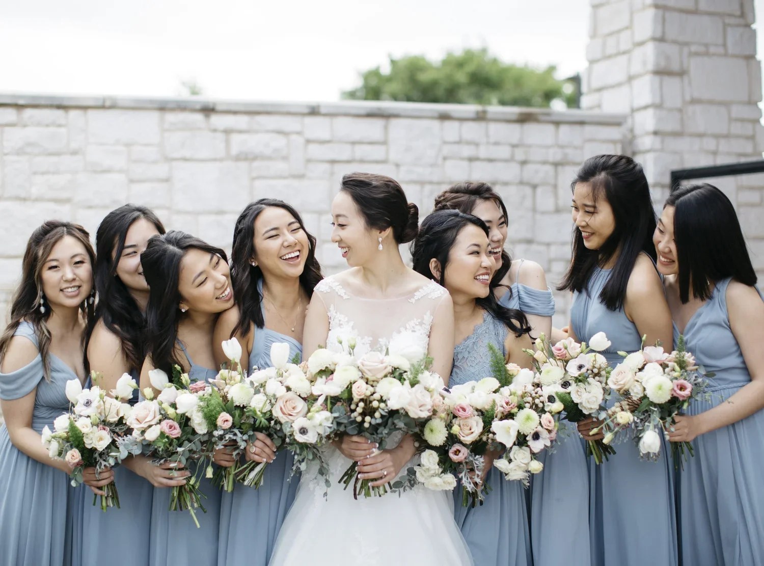A bride and her bridesmaids pose for a photo, all holding large bouquets of flowers. They are smiling and laughing together outdoors against a stone wall.