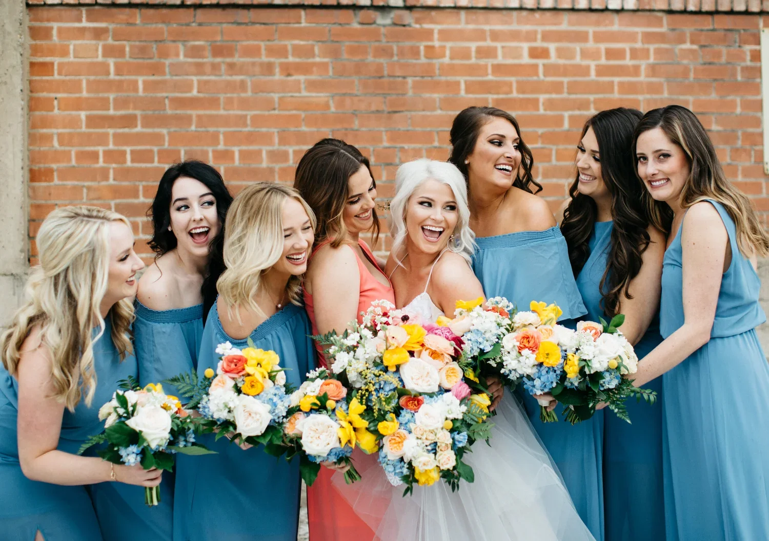 A group of women at a wedding, including the bride with white hair and a bride in a pink dress, surrounded by bridesmaids in blue dresses, holding bouquets of colorful flowers, standing in front of a brick wall.
