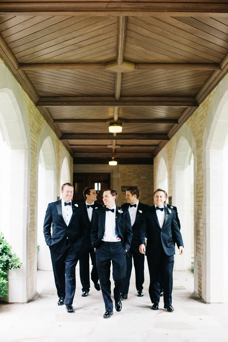 Six men in black tuxedos walking outdoors in a covered walkway with arches, smiling and looking at each other.