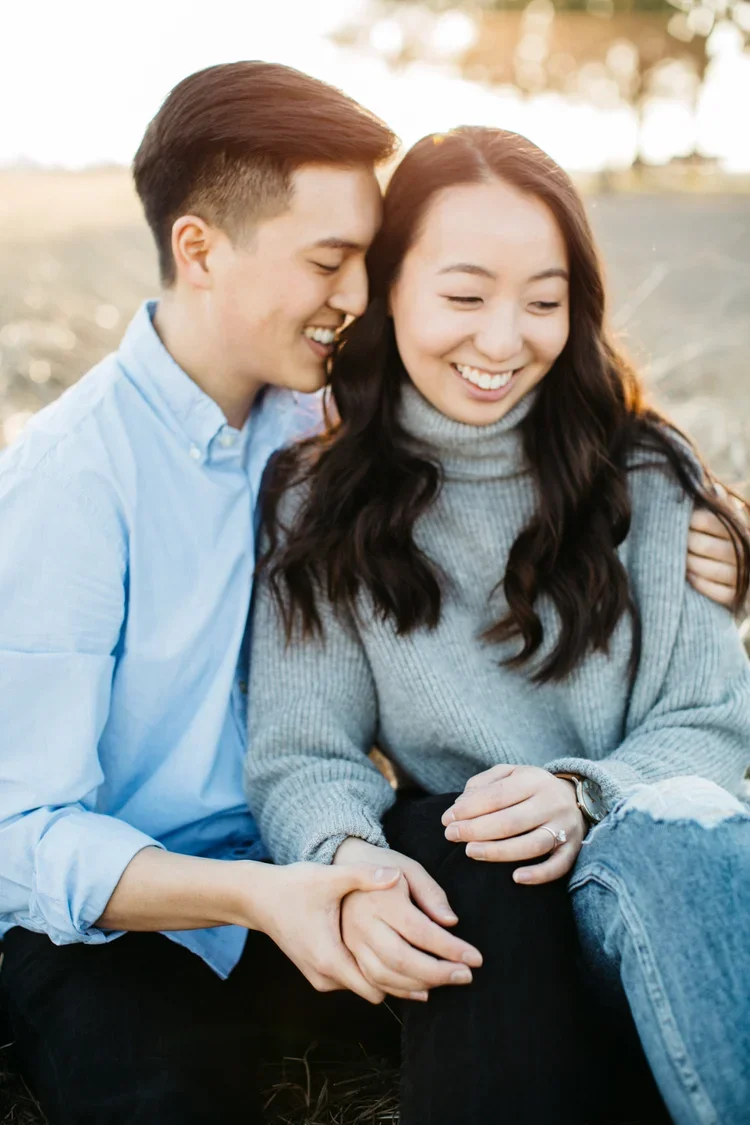 A smiling young man and woman sitting outdoors, the man whispering into the woman's ear while holding her hand, during sunset.