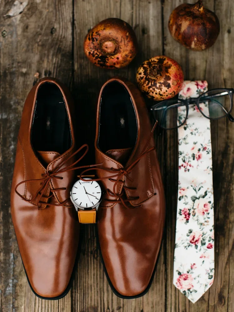 A pair of brown leather dress shoes, a wristwatch with a yellow strap, a pair of eyeglasses, a floral-patterned tie, and three ripe pomegranates on a wooden surface.