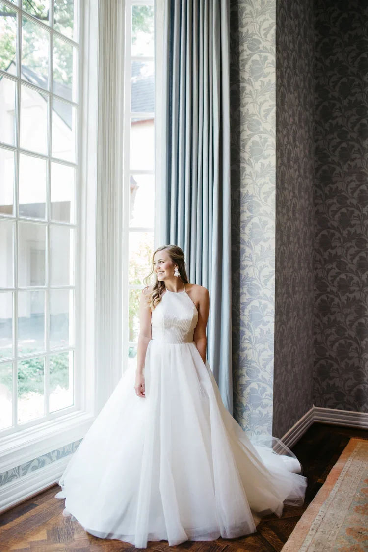A woman in a white wedding dress standing next to a large window with sheer curtains inside a decorated room.