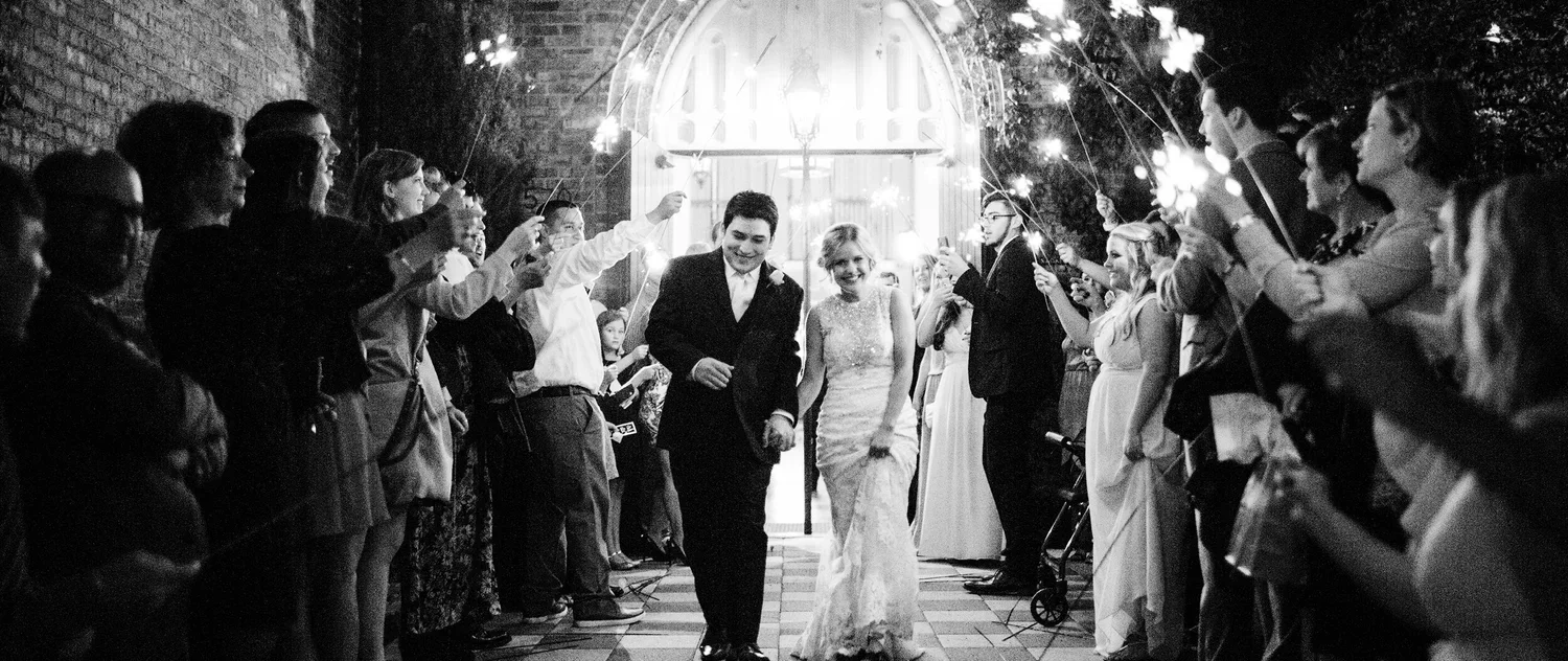 Black and white photo of a wedding celebration with a bride and groom walking through an aisle made by guests holding sparklers.
