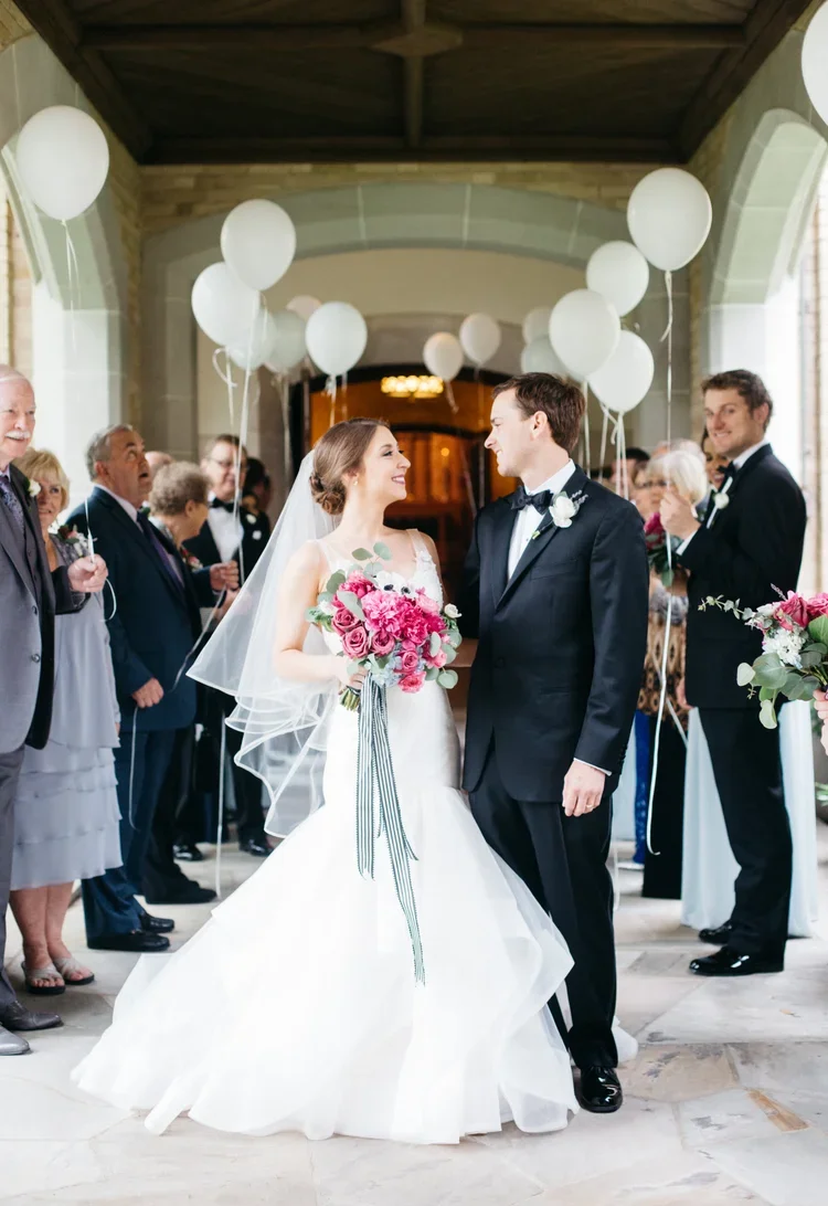A bride and groom hold hands and smile at each other during their wedding ceremony, surrounded by guests holding white balloons in an elegant indoor setting with large windows and a vaulted ceiling.