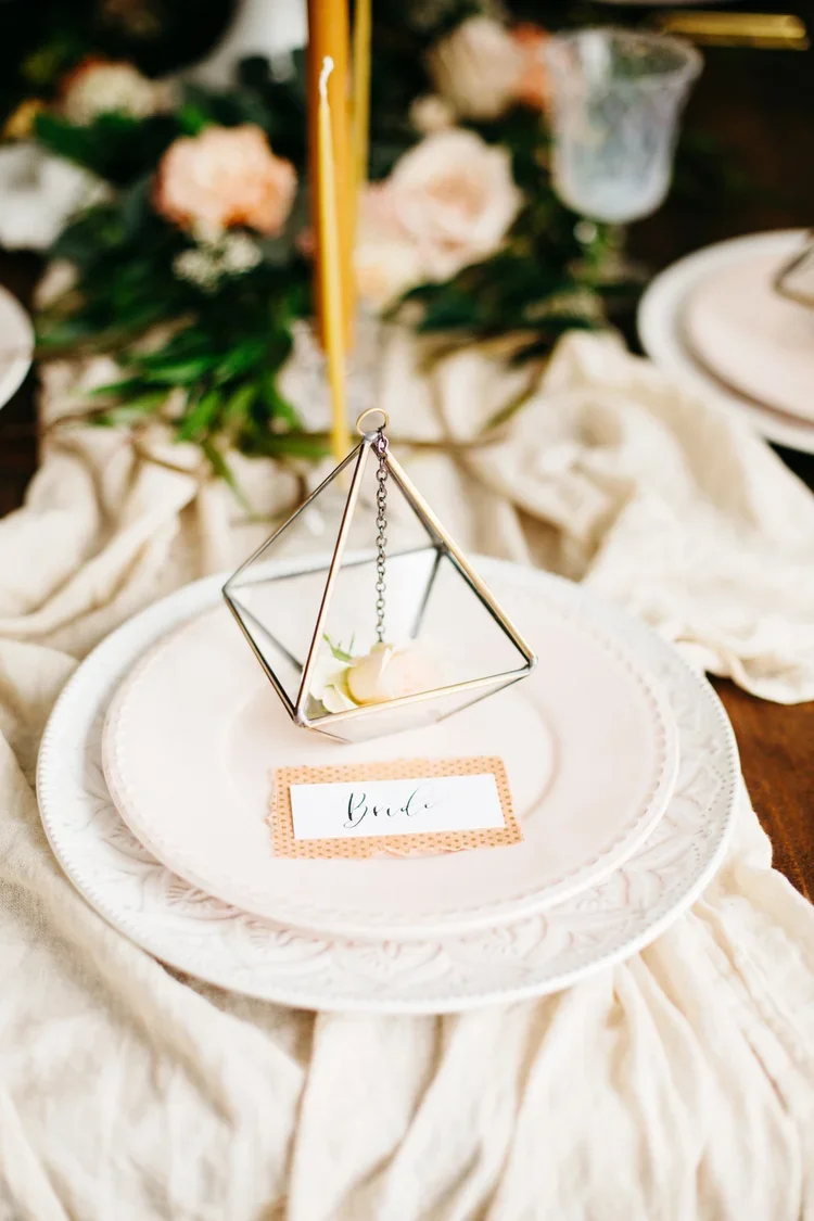 Elegant table setting with a geometric glass decoration on a white plate, a small flower inside the decoration, a 'bride' name tag, and floral arrangements in the background.