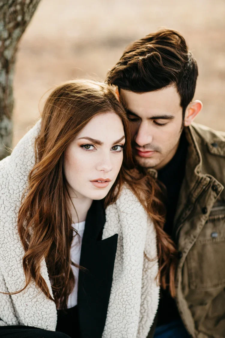 A young woman with long red hair and a young man with dark hair are sitting close together outdoors. The woman is looking directly at the camera, while the man has his eyes closed and his face is nearly touching her forehead. The background is blurre