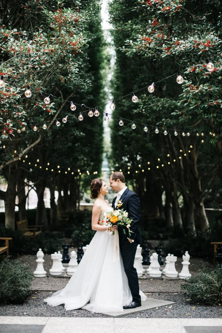Bride and groom standing close together under string lights in an outdoor garden, with the bride holding a bouquet of flowers.