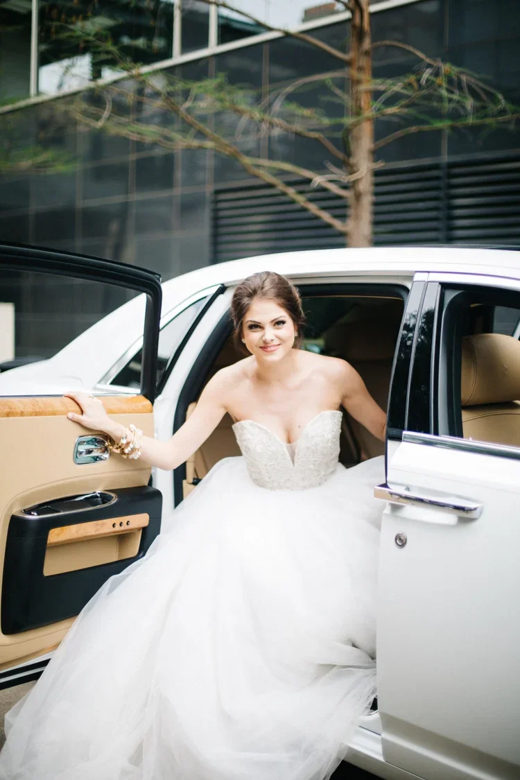 Bride in a white wedding dress stepping out of a luxury car with a beige interior, holding the door open with one hand, during daytime.