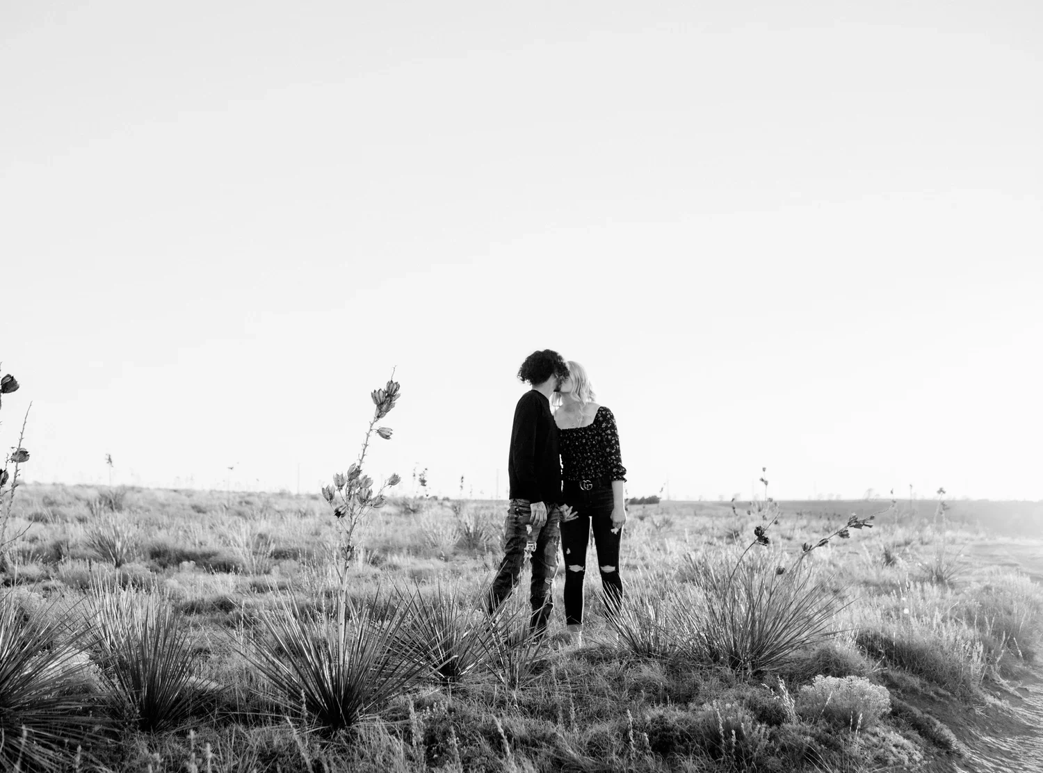 Two women stand close together in a vast open field with sparse desert plants, touching foreheads in a moment of intimacy, with a bright sky overhead.