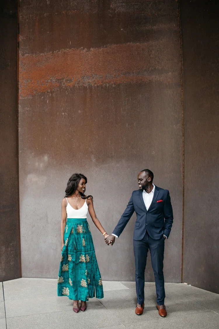 A couple holding hands and smiling at each other in front of a textured, rust-colored wall. The woman is wearing a white sleeveless top and a long turquoise skirt with gold patterns. The man is dressed in a navy blue suit with a white shirt and brown