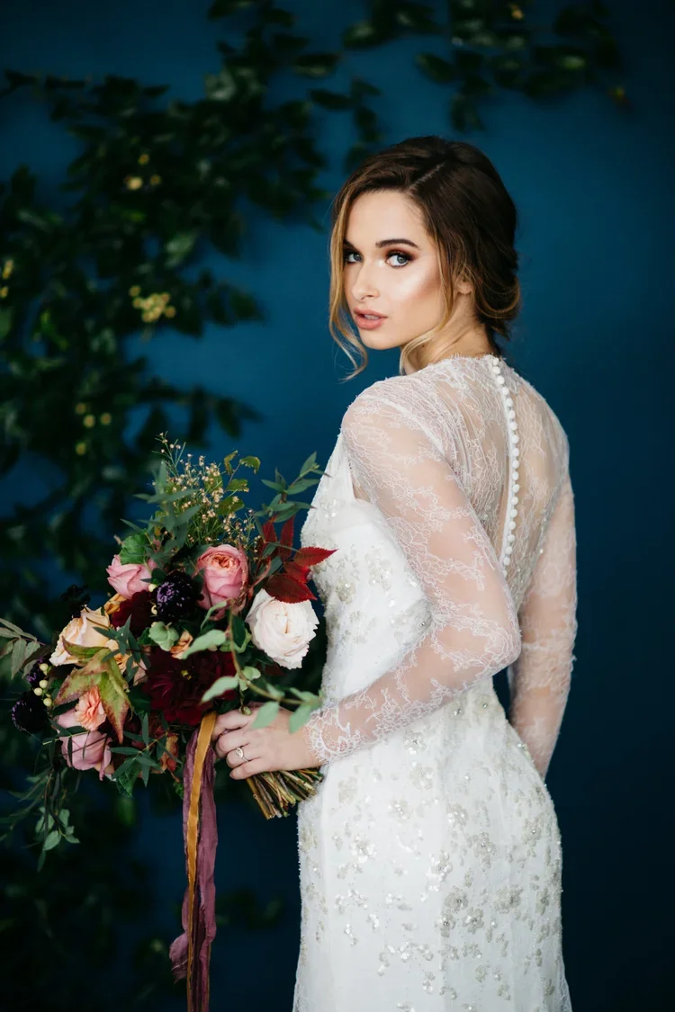 A woman in a white wedding dress holding a bouquet of pink, white, and dark purple flowers against a blue background with green foliage.
