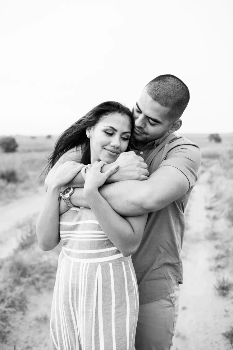 A black and white photo of a man and woman embracing outdoors. The man is hugging the woman from behind, and they look content and close to each other.