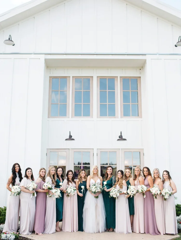 A bride and her bridesmaids pose in front of a white barn with large windows, all holding bouquets of white flowers.