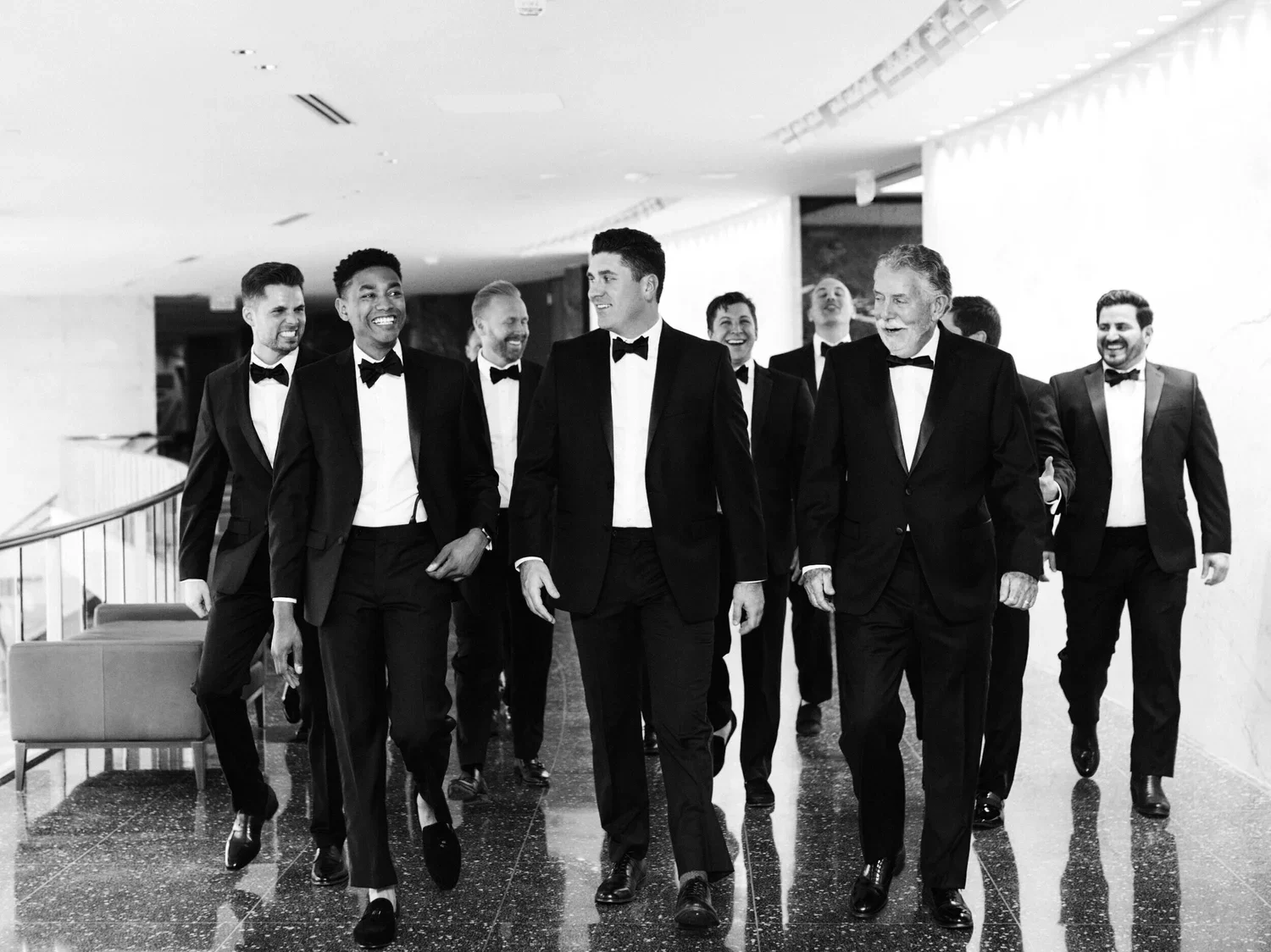 A group of men in tuxedos walking together in a modern indoor setting, smiling and engaging with each other.