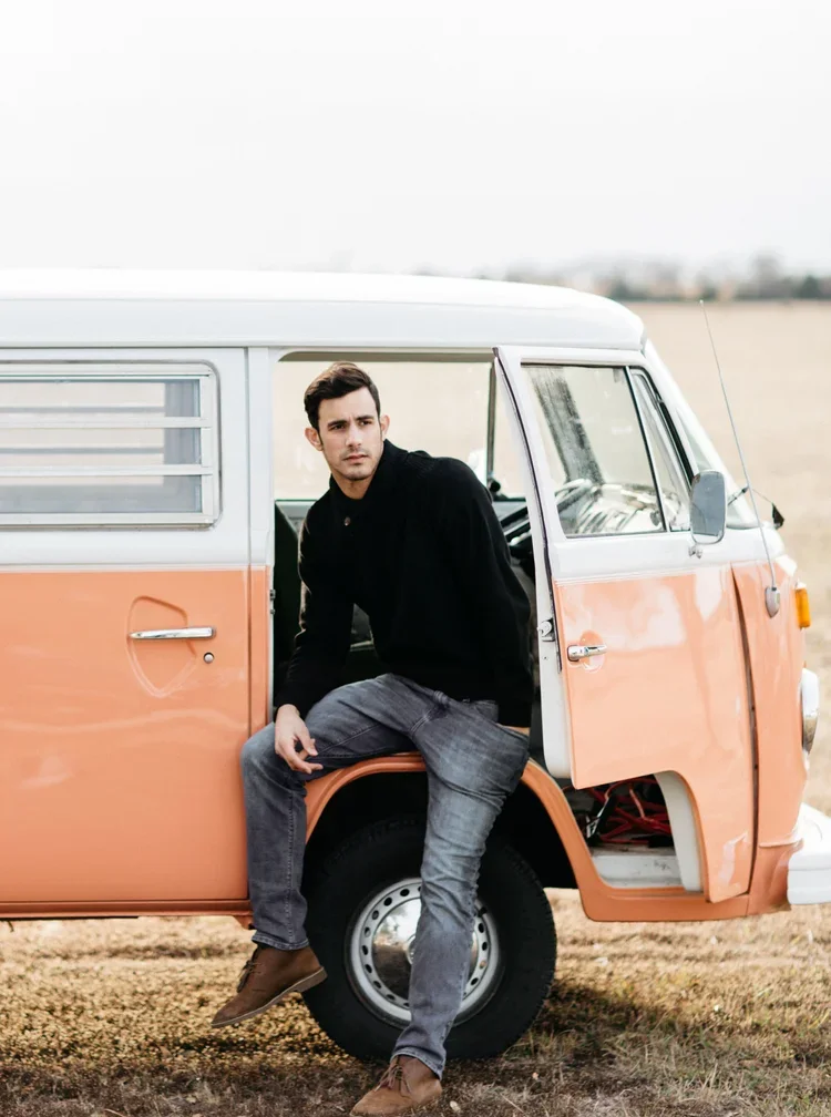 A young man sitting on the open door of an orange and white vintage van, parked in a field with a blurred landscape in the background.