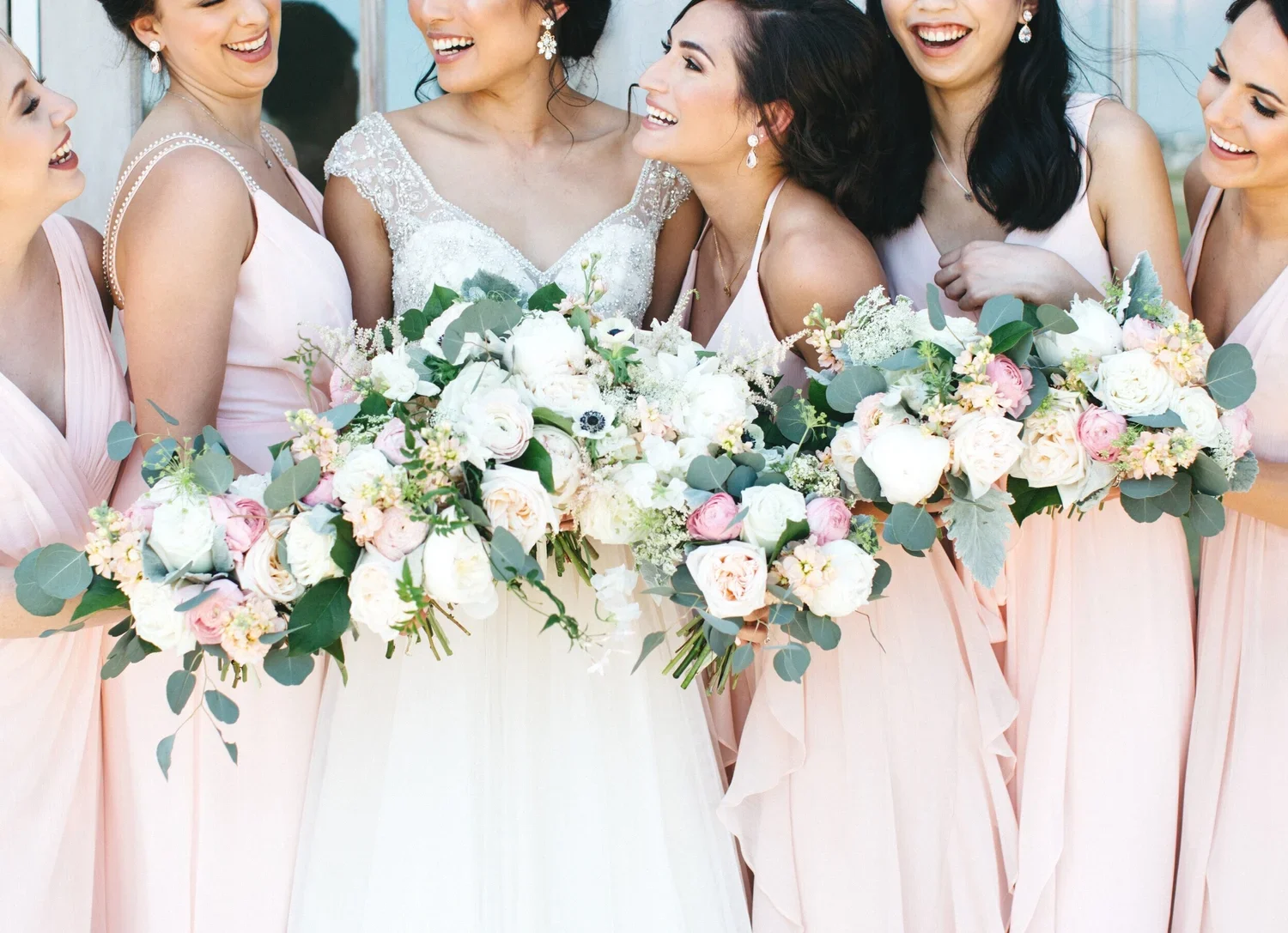 Group of women, including a bride and bridesmaids, smiling and dressed in wedding attire, holding large floral bouquets.