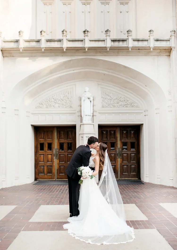 A bride and groom sharing a kiss in front of a church entrance, with the bride holding a bouquet of flowers and wearing a white wedding dress with a veil.