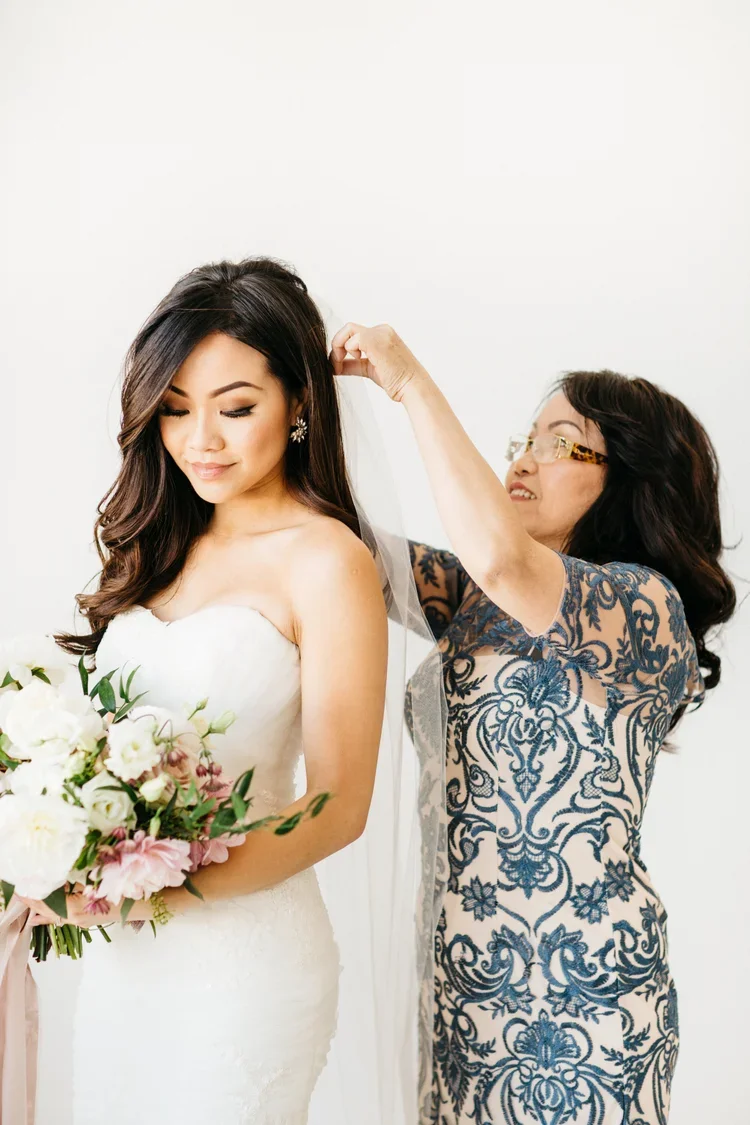 A bride with long dark hair in a white strapless wedding dress holding a bouquet of white and pink flowers, while a woman with glasses in a dark blue and beige lace dress adjusts her veil.