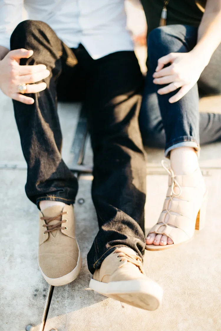 Close-up of a person sitting on a bench, wearing black jeans and tan shoes, with another person kneeling beside them, holding their leg.
