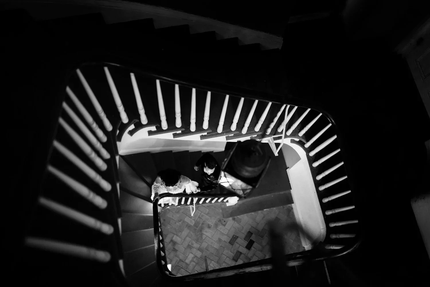 Top-down view of a spiral staircase with two people walking up, one wearing white and one wearing dark clothing.