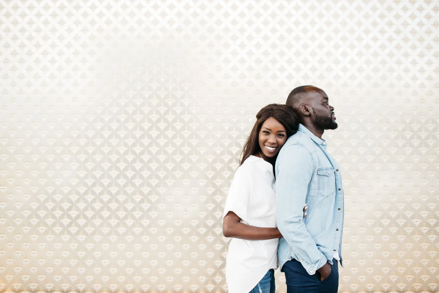 A smiling Black woman stands behind a serious Black man, hugging him from behind, against a light patterned background.