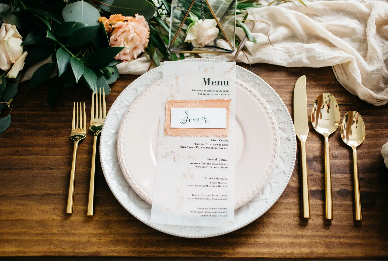 Elegant wedding table setting with a white plate, gold flatware, and a menu card for Caroline and Theodore Roberts' wedding on April twelfth, featuring a floral and greenery centerpiece, and a "Groom" label on the menu.