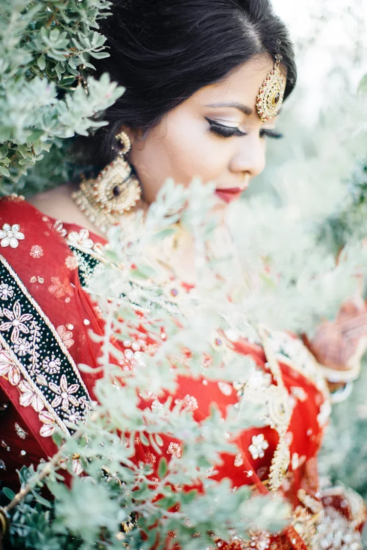 A woman dressed in traditional South Asian red and black bridal attire with detailed embroidery, wearing elaborate gold and gemstone jewelry, and having long black hair. She is surrounded by greenery and appears to be in a contemplative mood.