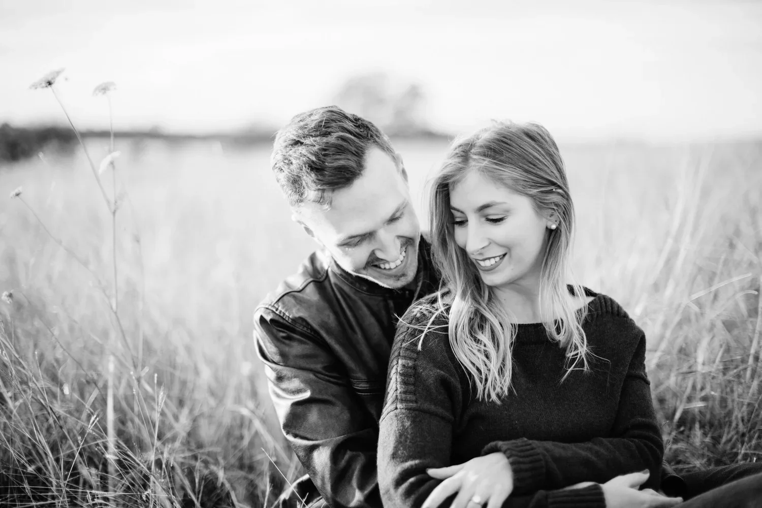A black and white photo of a smiling couple sitting in a grassy field, close together, enjoying each other's company.