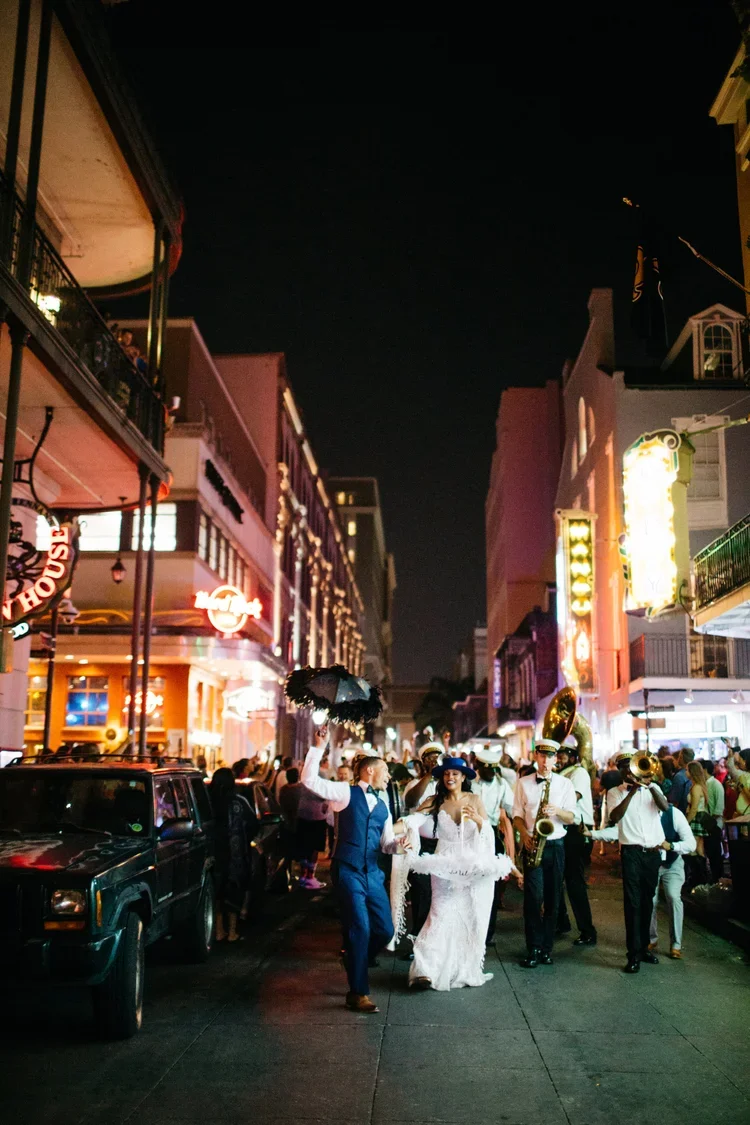 Nighttime street scene with a parade featuring a bride and groom, dressed in wedding attire, walking beside a jazz band playing music. The street is lined with illuminated buildings and signs, with onlookers watching from the sides.