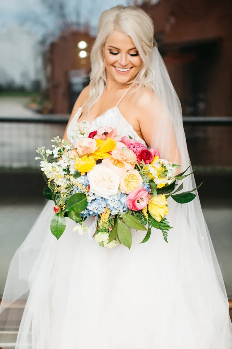 A smiling bride with platinum blonde hair holding a colorful bouquet of flowers, wearing a white wedding dress and veil, outdoors with a blurred background.