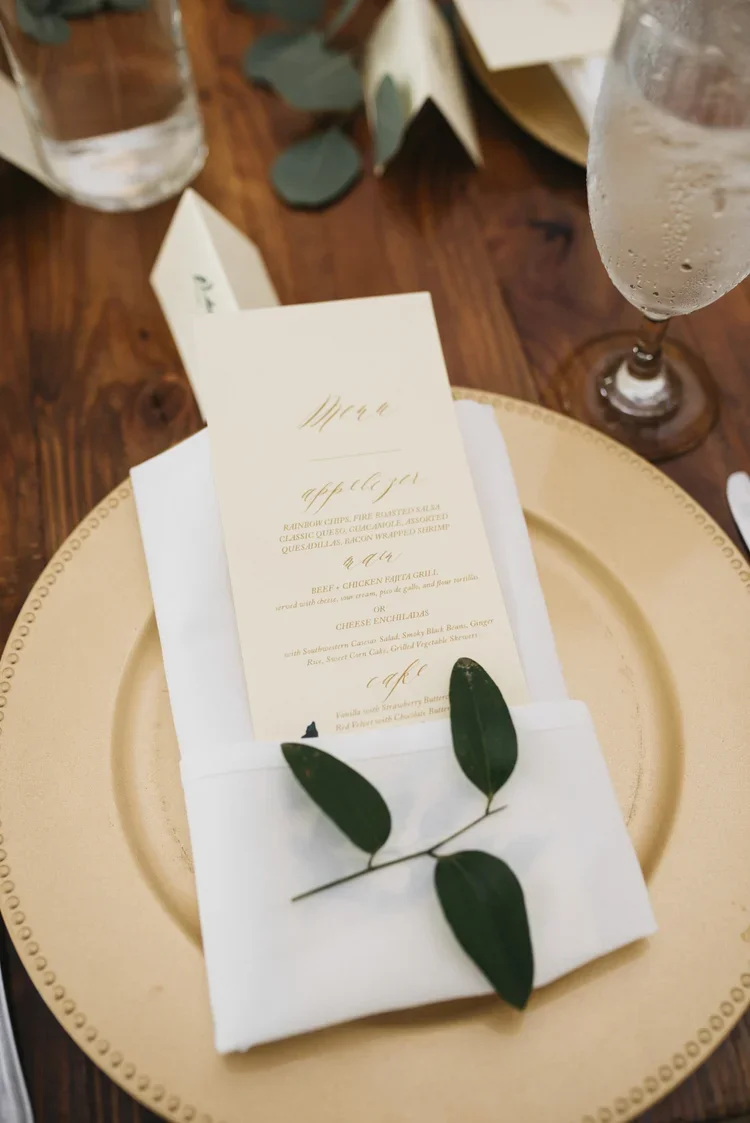 A place setting with a gold charger plate, a neatly folded white napkin with a green leafy branch, and a printed menu placed on top. The menu features elegant script and lists various food options.