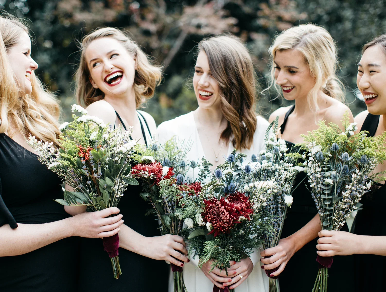 A group of women in black dresses holding bouquets of flowers, laughing and chatting outdoors during a celebration.