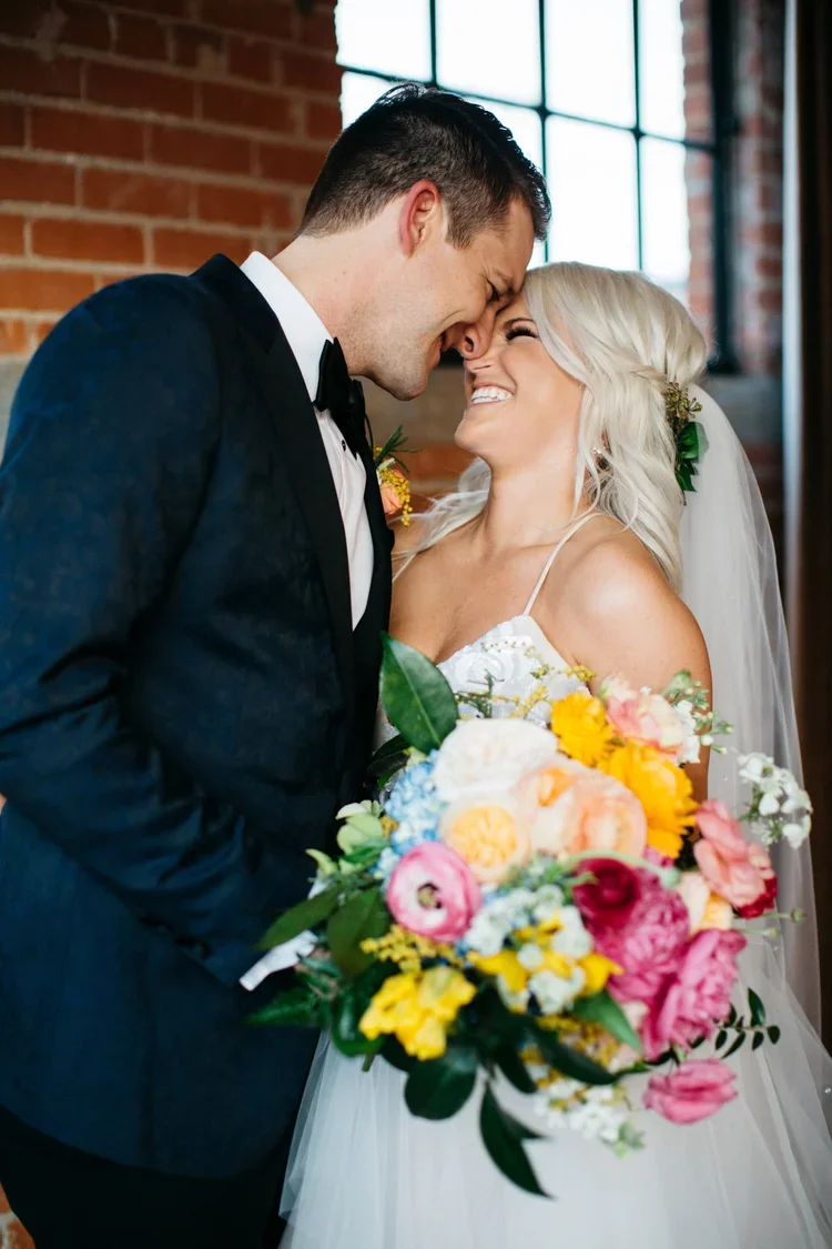 Bride and groom in an indoor wedding, close together with foreheads touching, smiling, holding a colorful bouquet of flowers, with a brick wall and window in the background.
