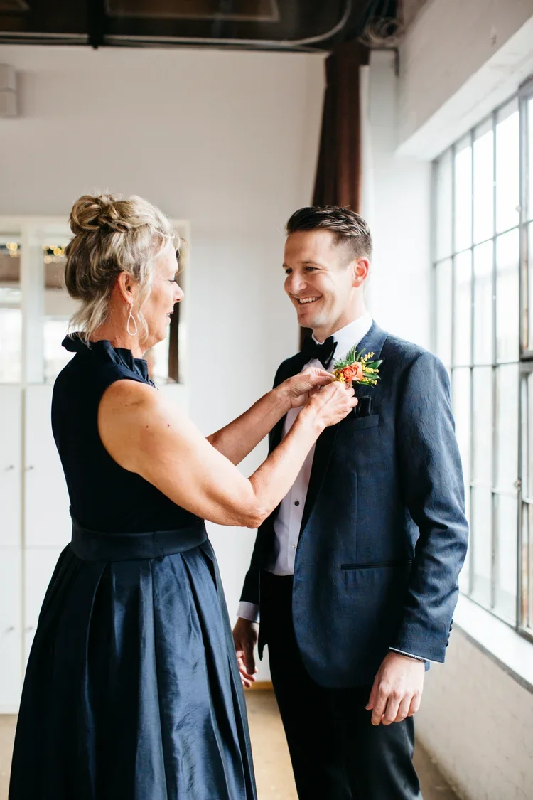 A woman pinning a boutonniere on a man's tuxedo at a wedding or formal event, standing near a large window.