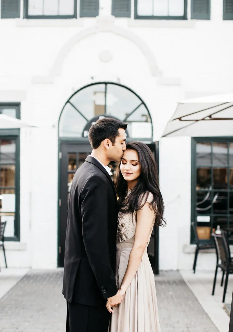 A man in a black suit kissing a woman in a beige dress on the forehead outdoors, with a white building and patio chairs in the background.