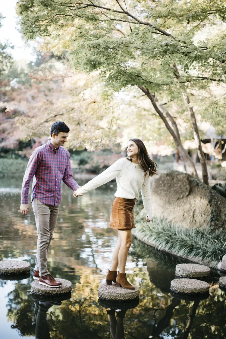 A young man and woman holding hands while standing on stones in a pond in a park with trees and fall foliage.
