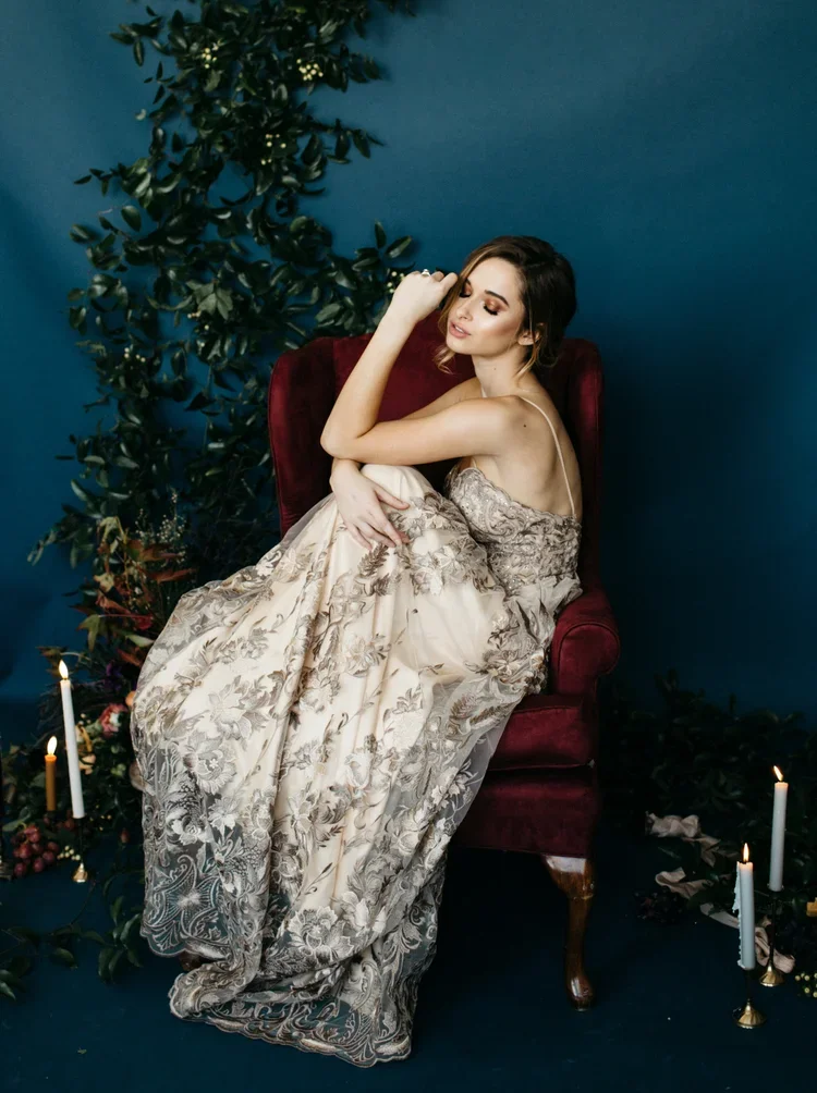 A woman in an elegant floral dress sits on a red velvet armchair, surrounded by candles and greenery against a blue wall.